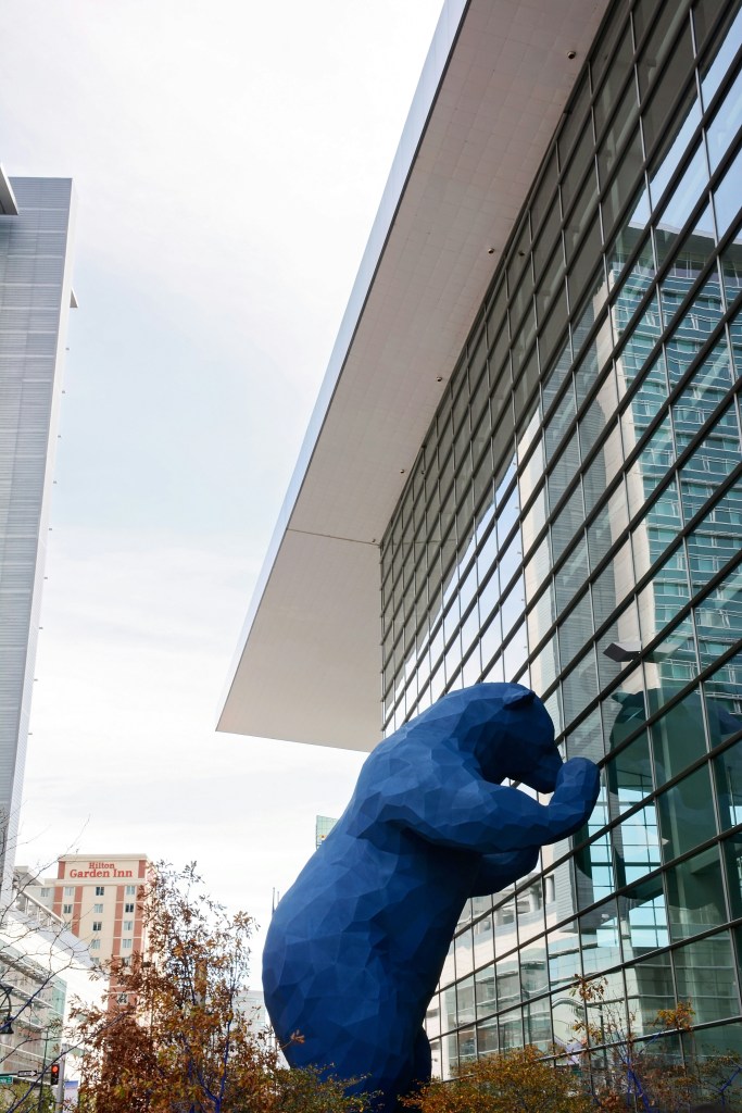 Image of a large, multi-story high blue bear sculpture, standing upright on its hind two legs and peering into the Denver Convention Center.